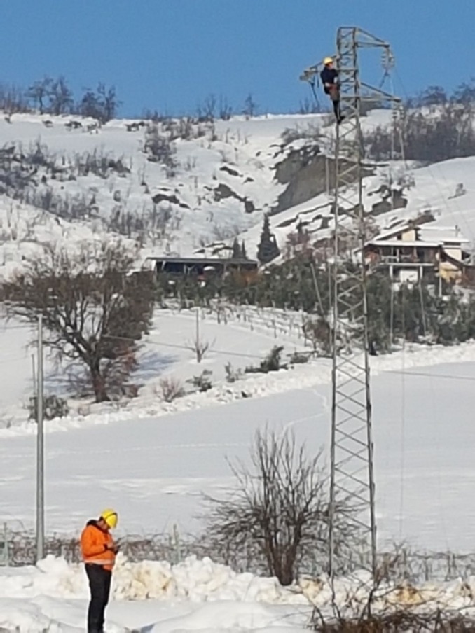 Operatore in quota su traliccio elettrico in paesaggio innevato, con collega a terra in sorveglianza